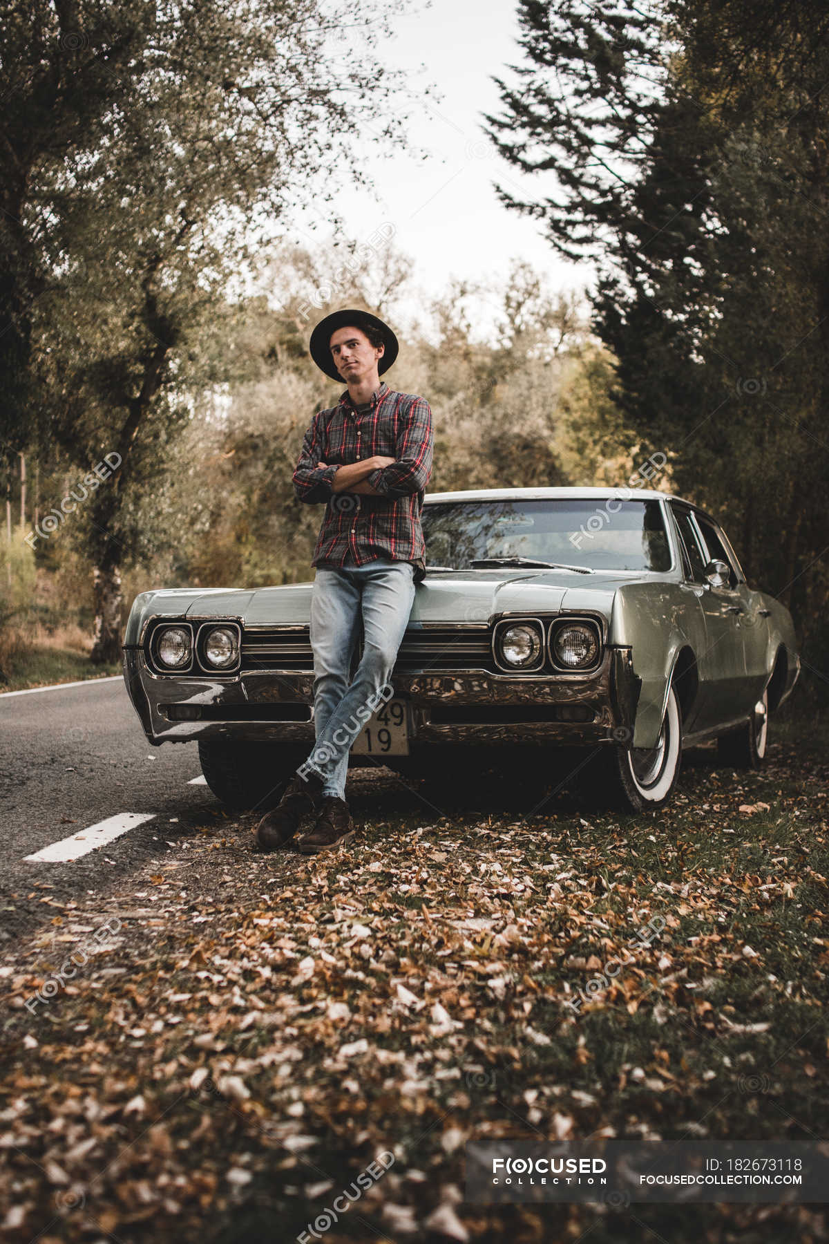 Confident young man with crossed arms leaning on trunk of retro car ...