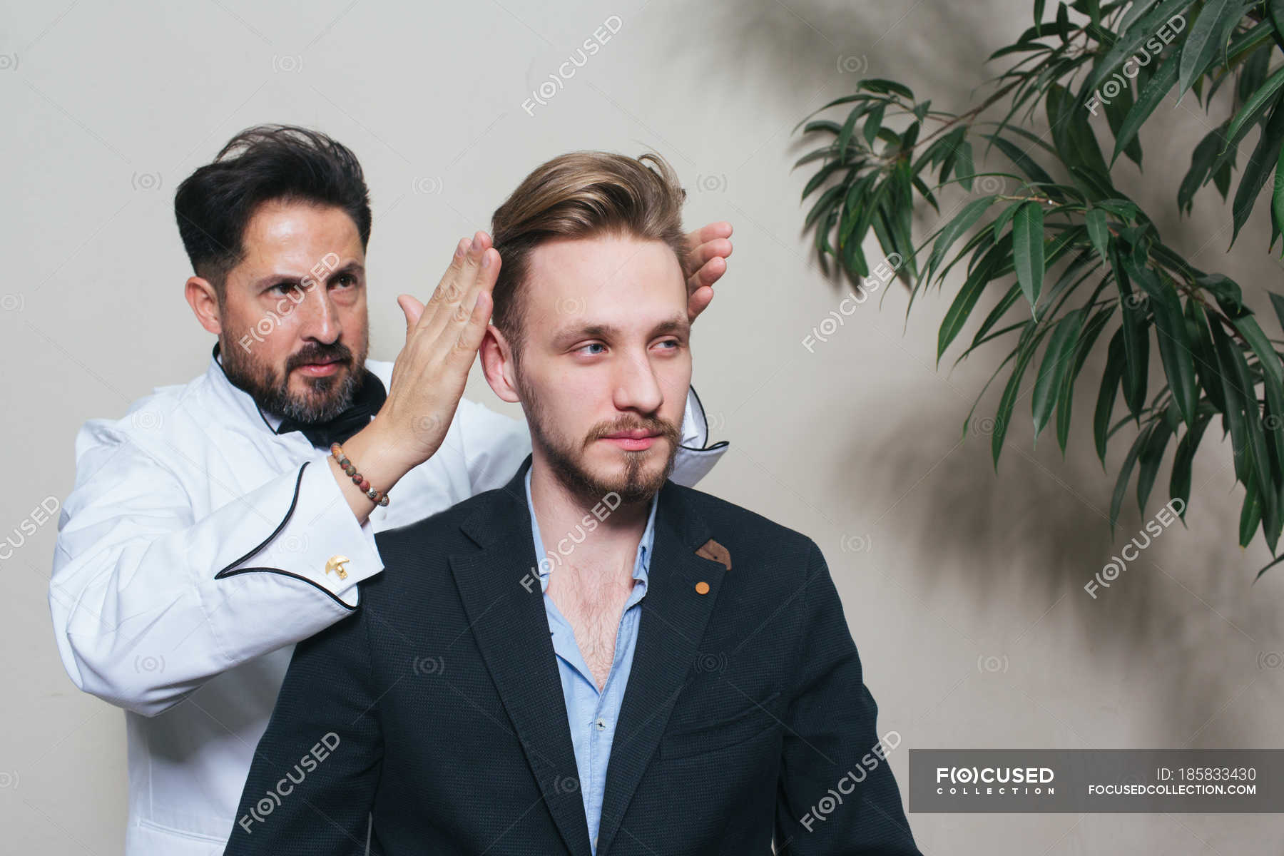 Barber Styling Hair Of Young Man In Suit In Hairdressing Salon