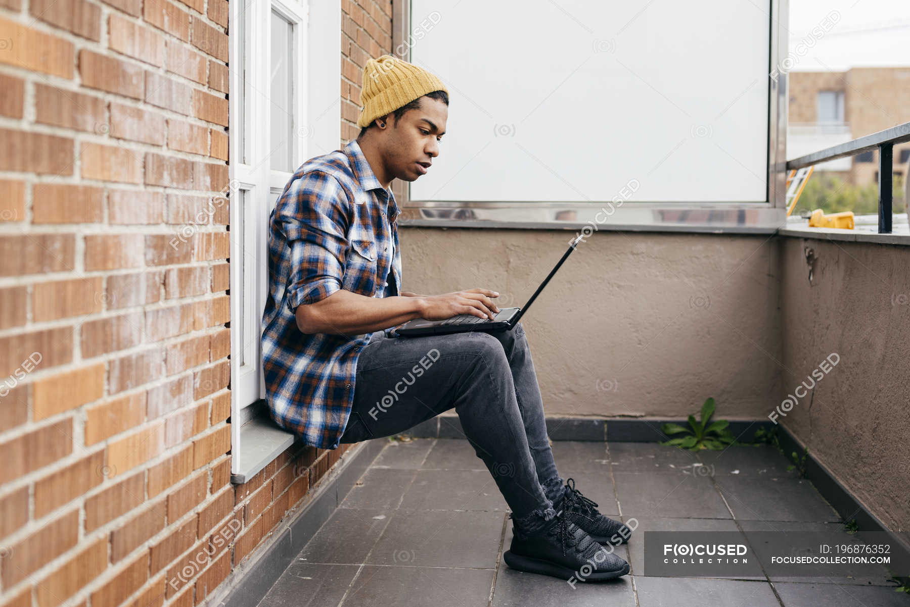 Side view of stylish man sitting on balcony and typing on laptop ...