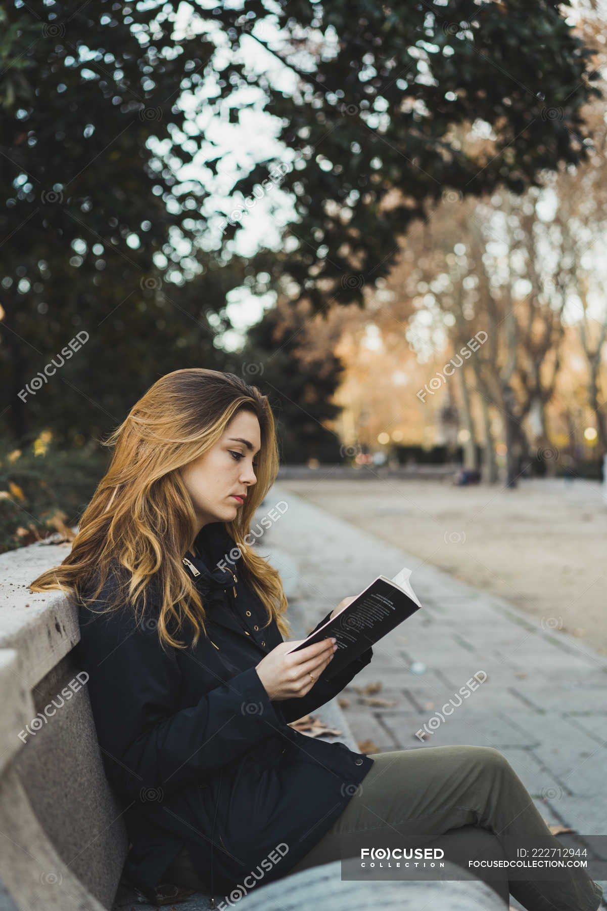 Young woman sitting on park bench and reading book — side view ...