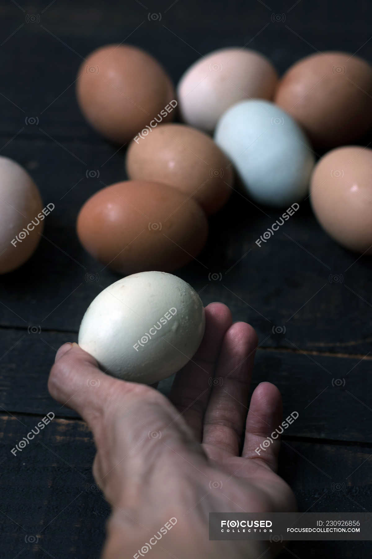 Human hand holding white egg above wooden table with heap of fresh