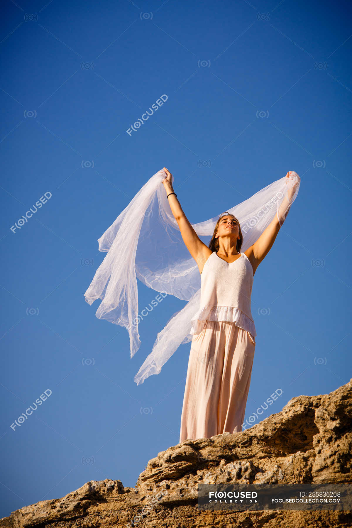 from-below-young-mysterious-woman-with-upped-hands-holding-white-textile-and-posing-on-rocks-and