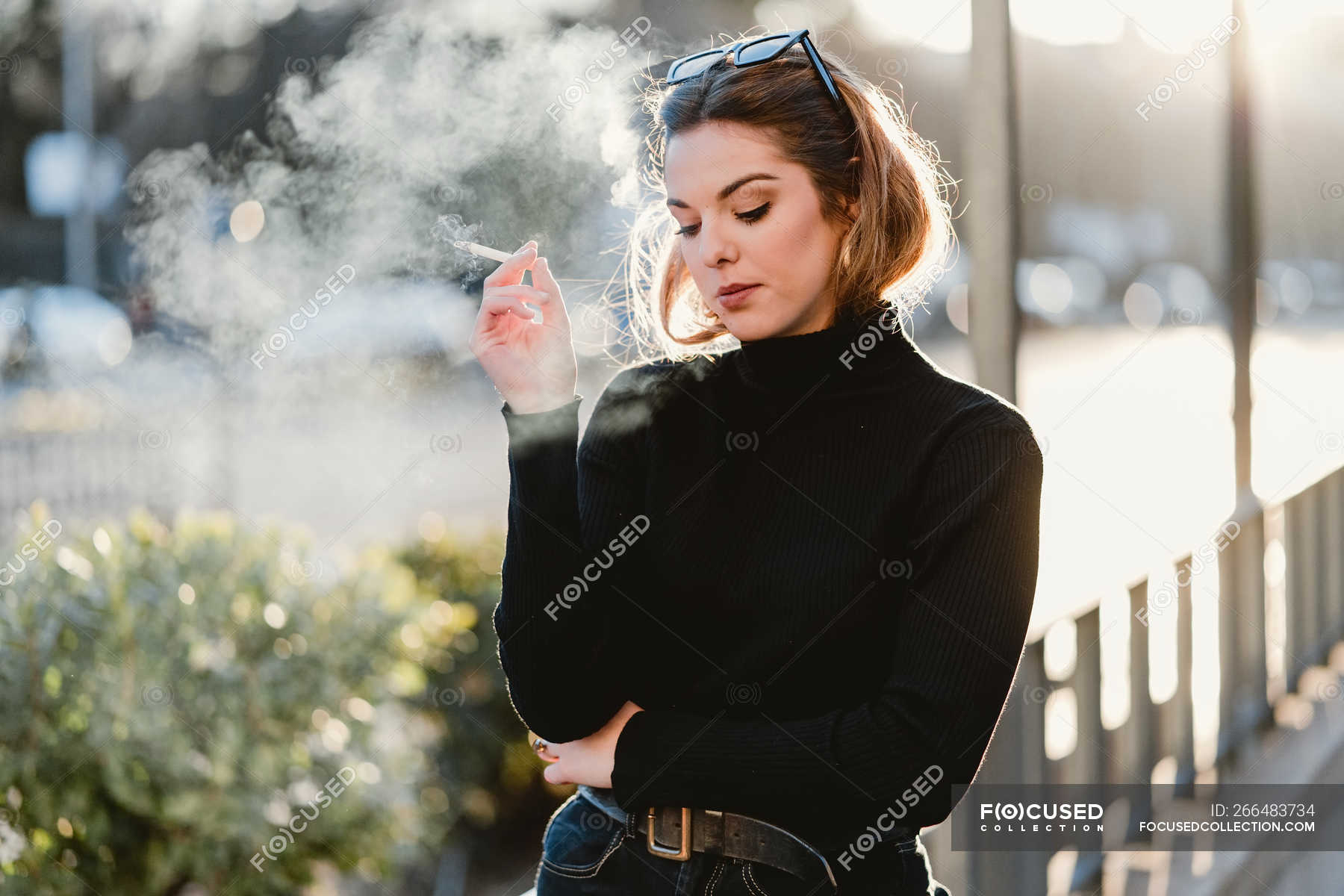 Young lady in trendy outfit exhaling fume while smoking cigarette on ...