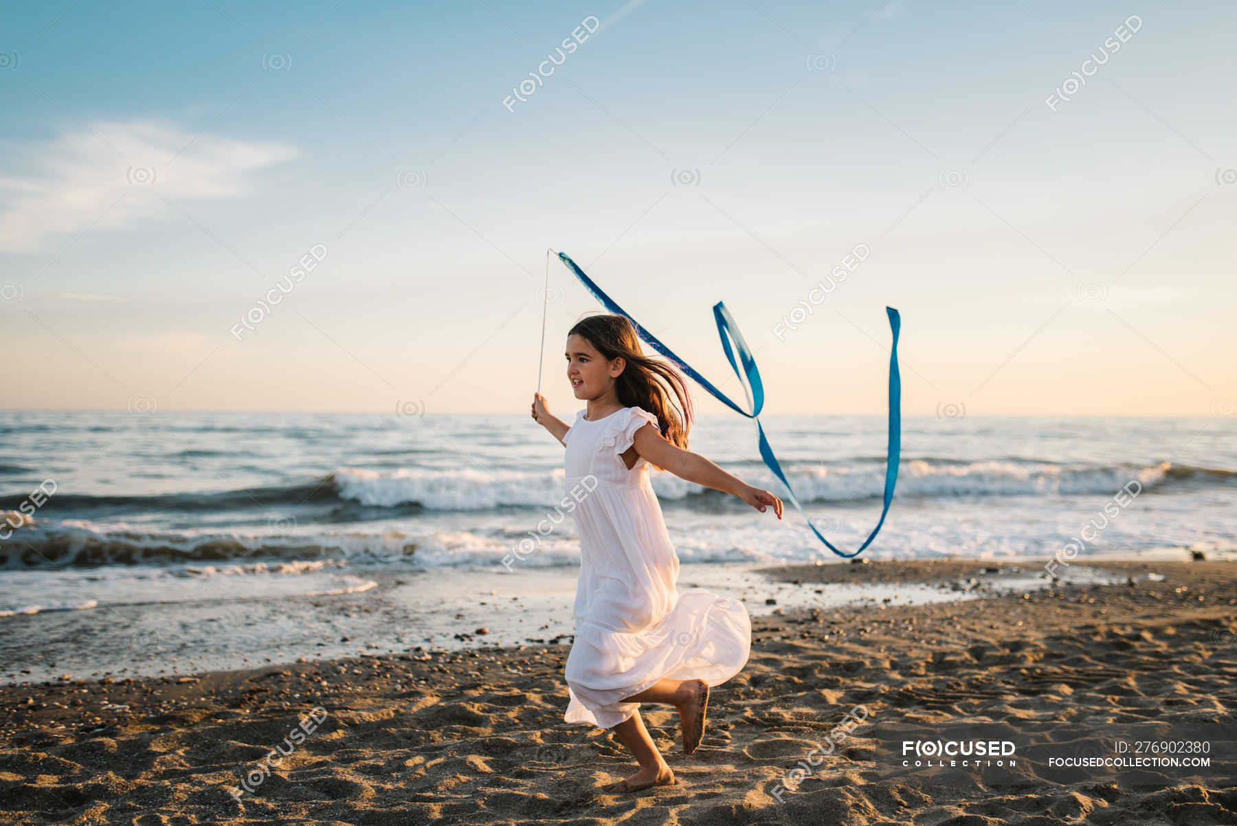 Little girl in white dress running along seashore and holding long blue band on background of