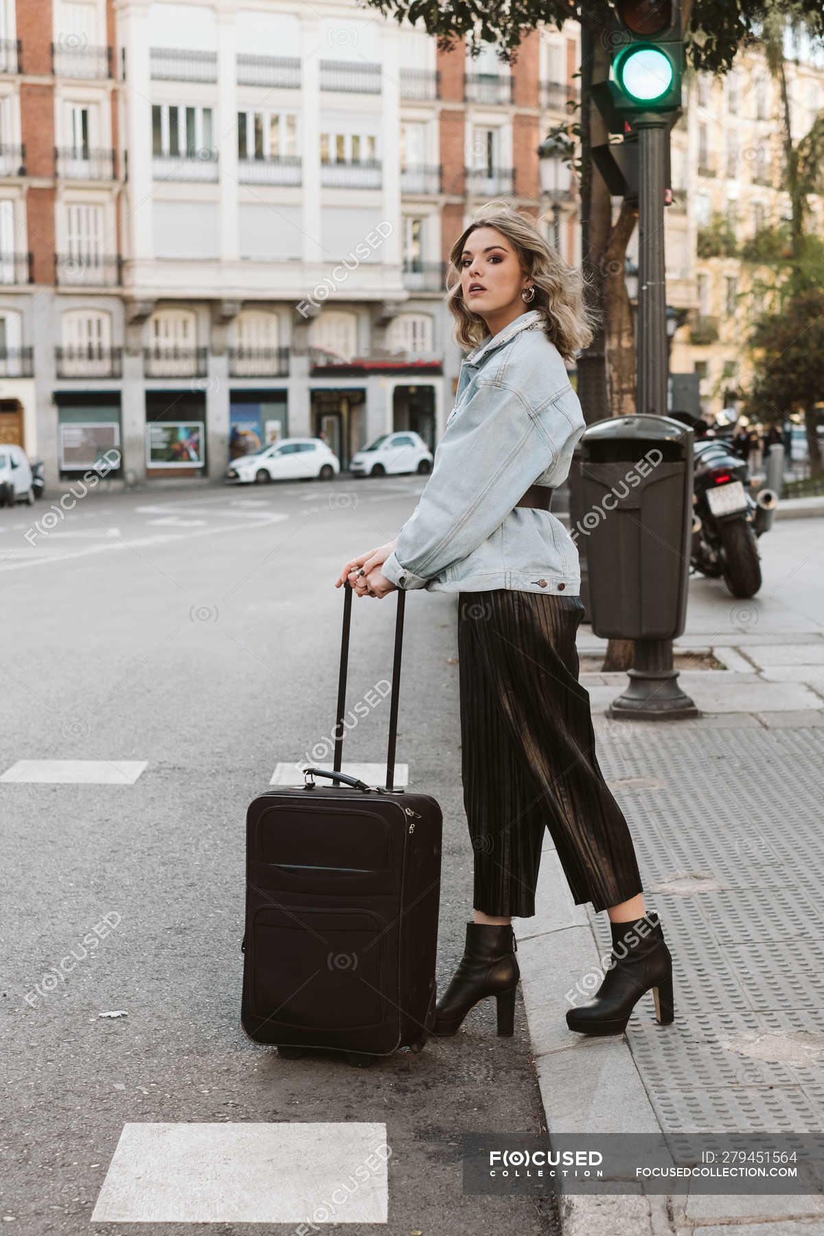 Stylish young woman with suitcase looking away while crossing city ...