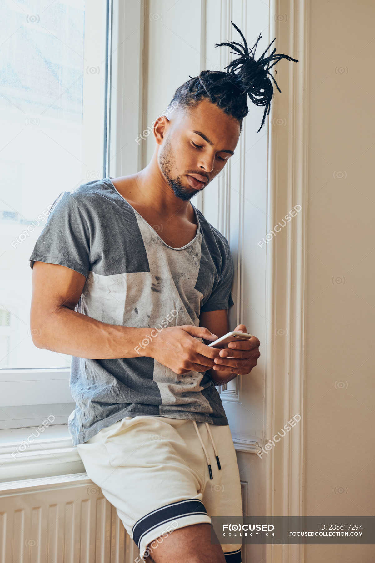 Young African American man with creative hairstyle leaning on windowsill at  home using mobile phone — gadget, subculture - Stock Photo | #285617294