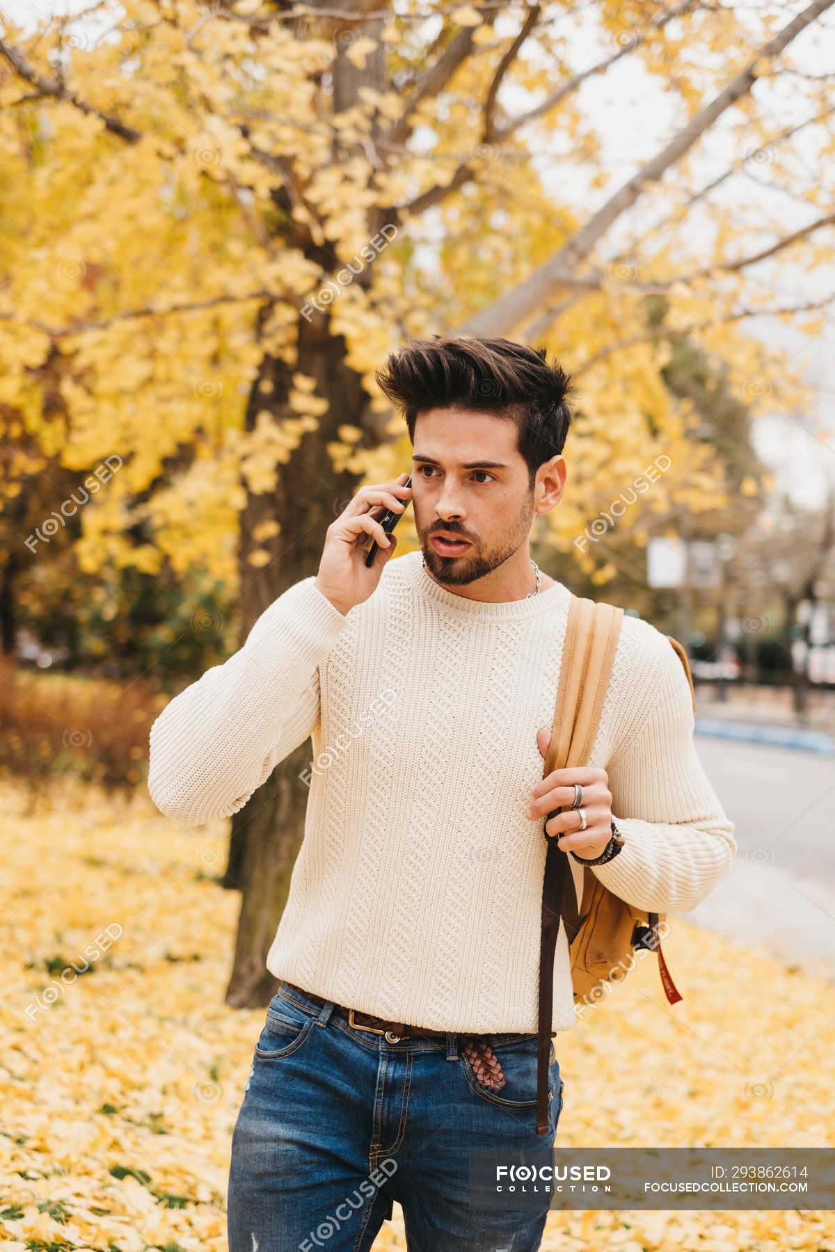 Handsome young man with trendy hairstyle walking on autumn leaves and  speaking on smartphone on daytime — communication, sweater - Stock Photo |  #293862614