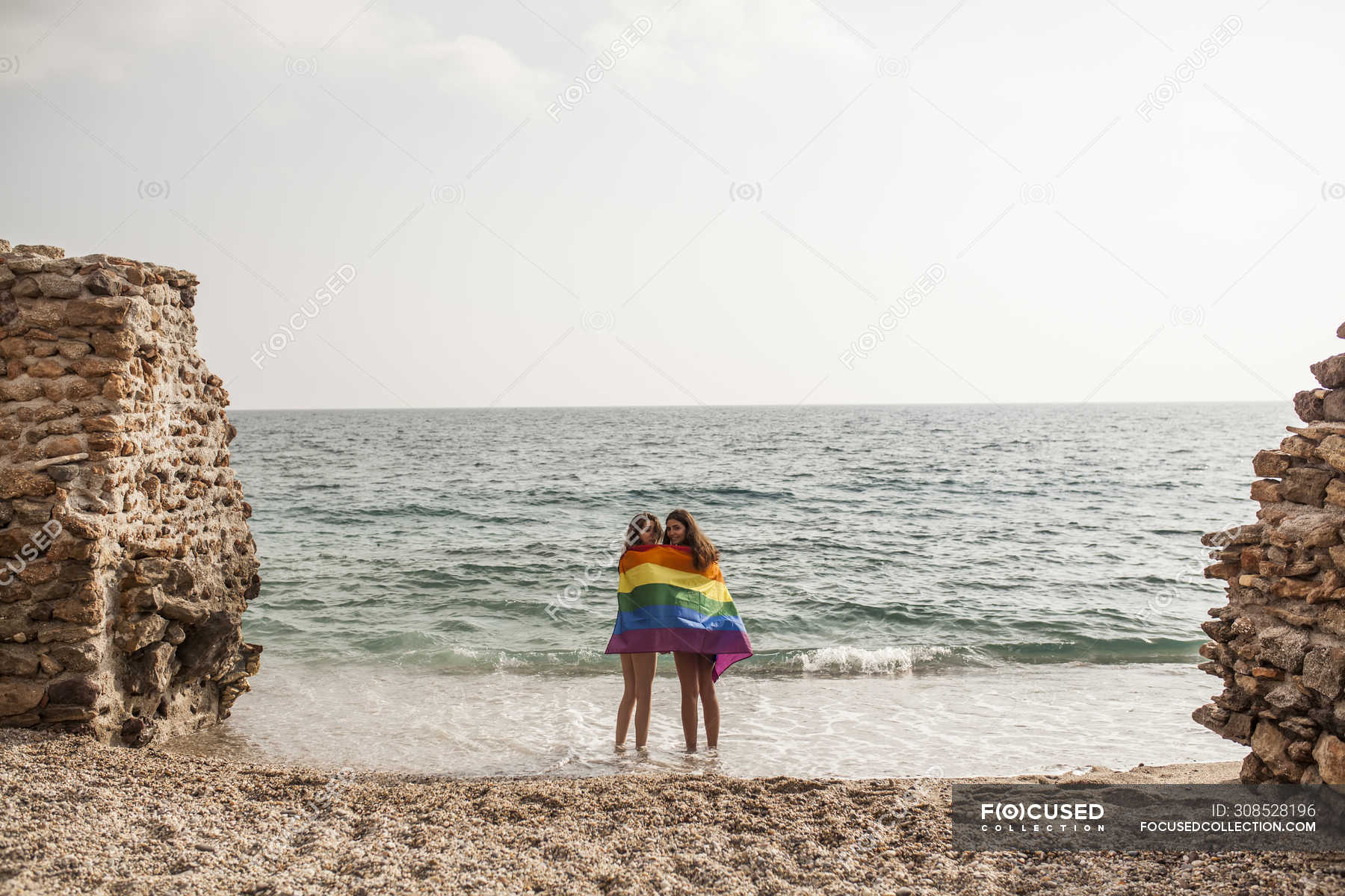 Back View Of Lesbian Couple Standing On Beach With Colorful Flag Of