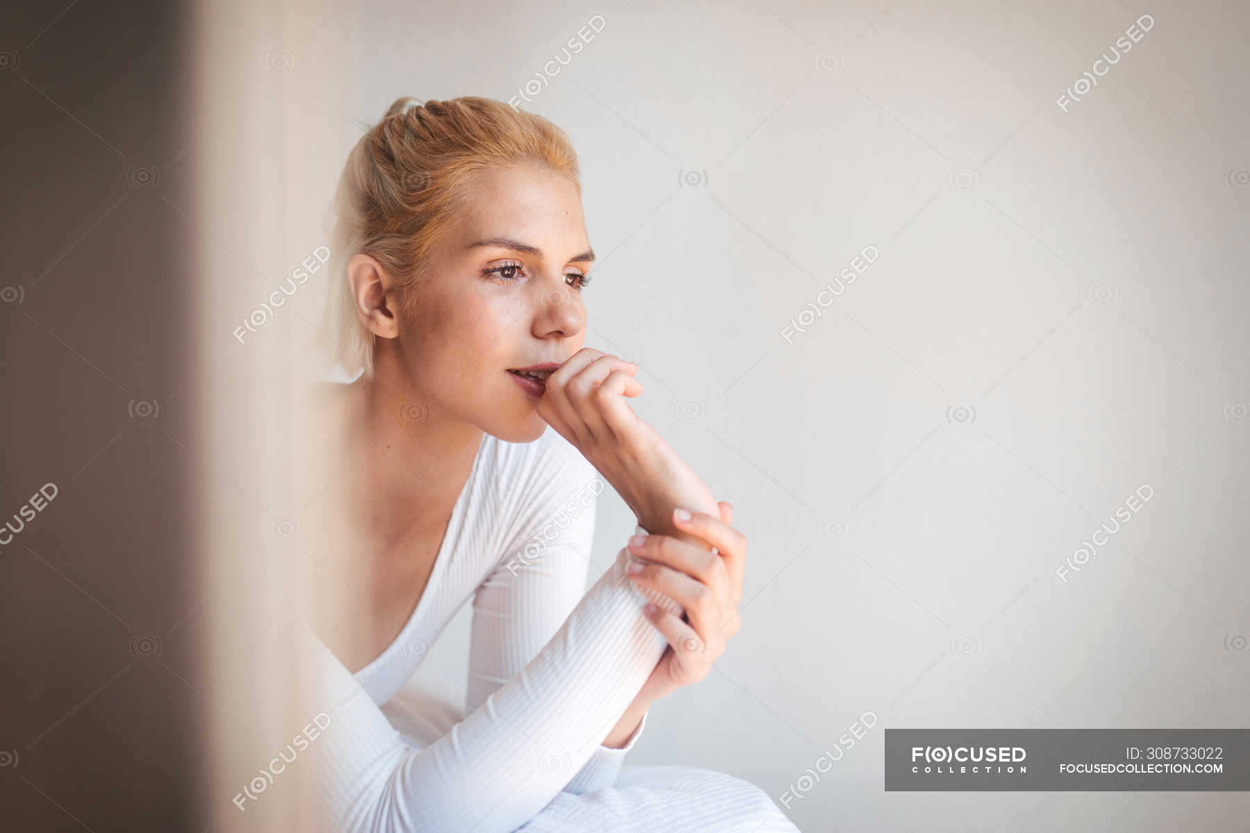 Young Woman With Blond Hair And In Bodysuit Looking Away While Sitting On Soft Bed Against White