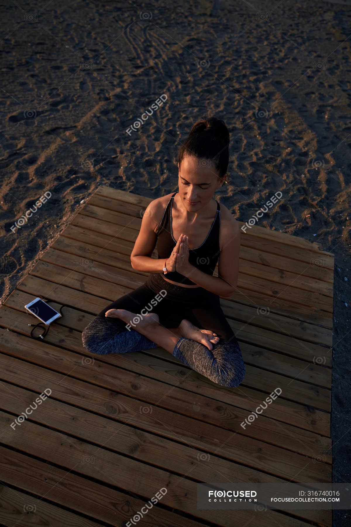 From Above Barefoot Woman In Sportswear Sitting In Lotus Pose On Lumber Path Near Sneakers And Smartphone And Meditating Summer Shore Stock Photo