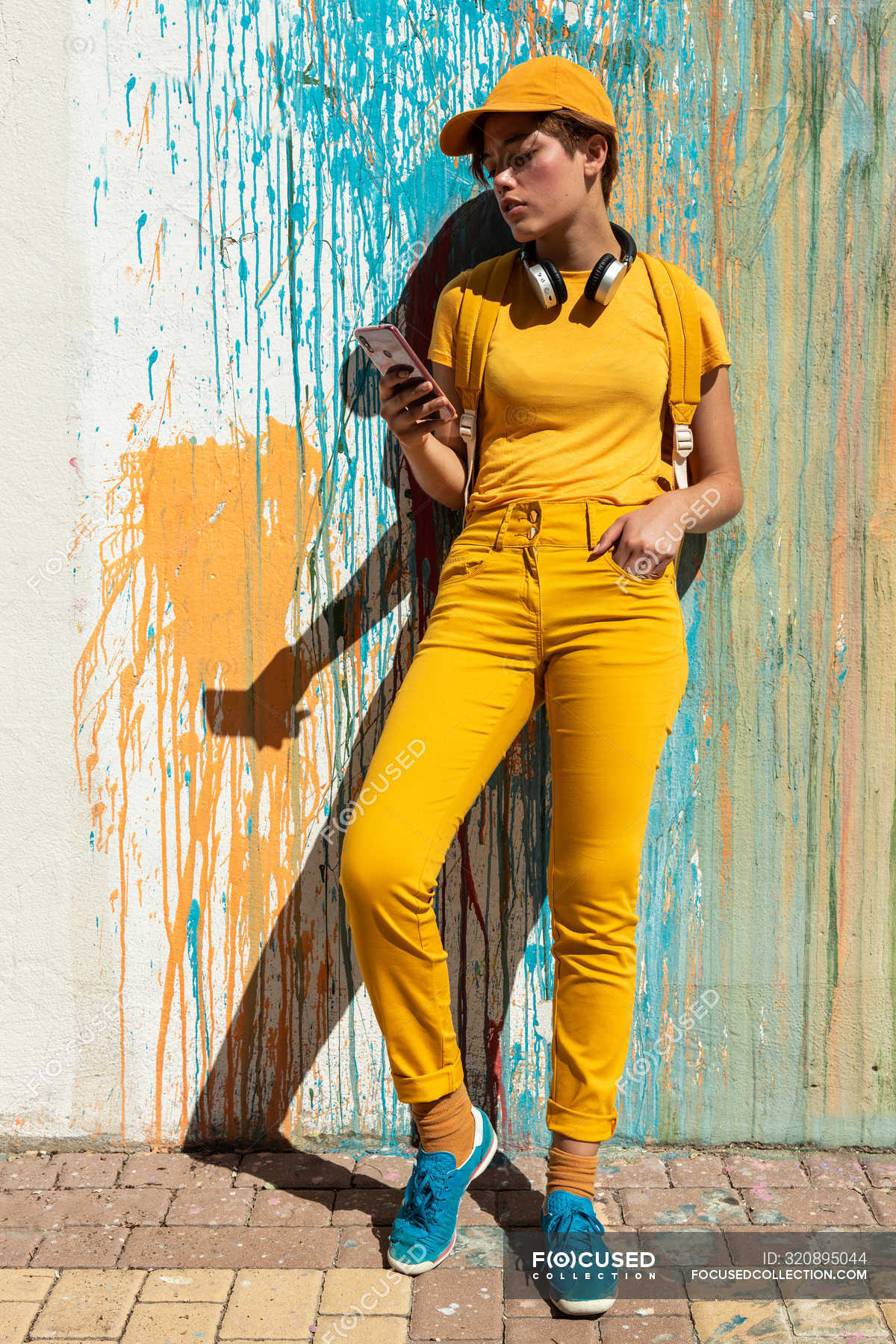 Stylish millennial in bright yellow outfit with headphones and smartphone  standing against wall with colorful paint drips — grunge, idea - Stock  Photo | #320895044