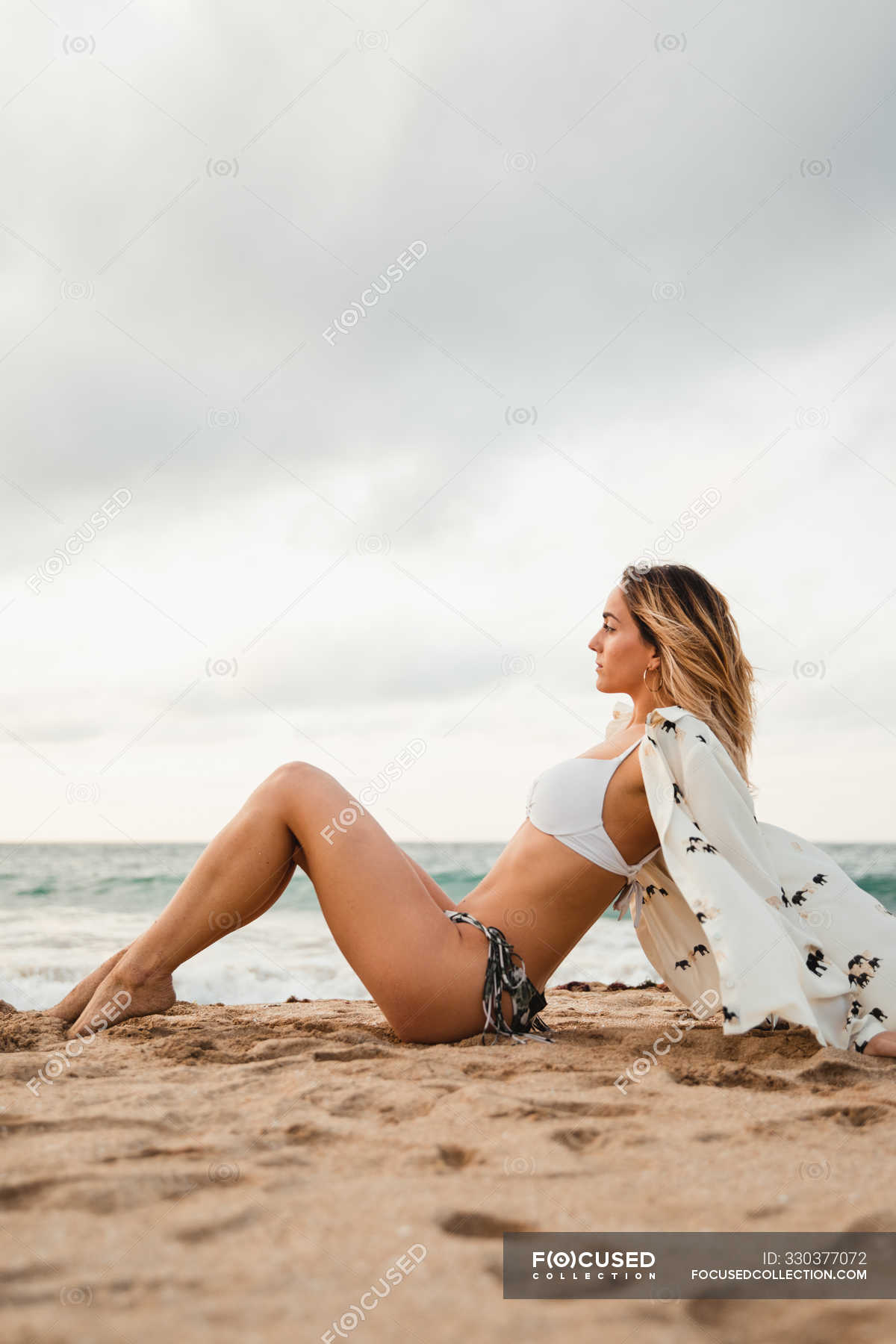 view of slim woman bikini and blouse leaning back and looking away while sitting on sandy coast against gray overcast sky — attractive, calm - Stock Photo | #330377072
