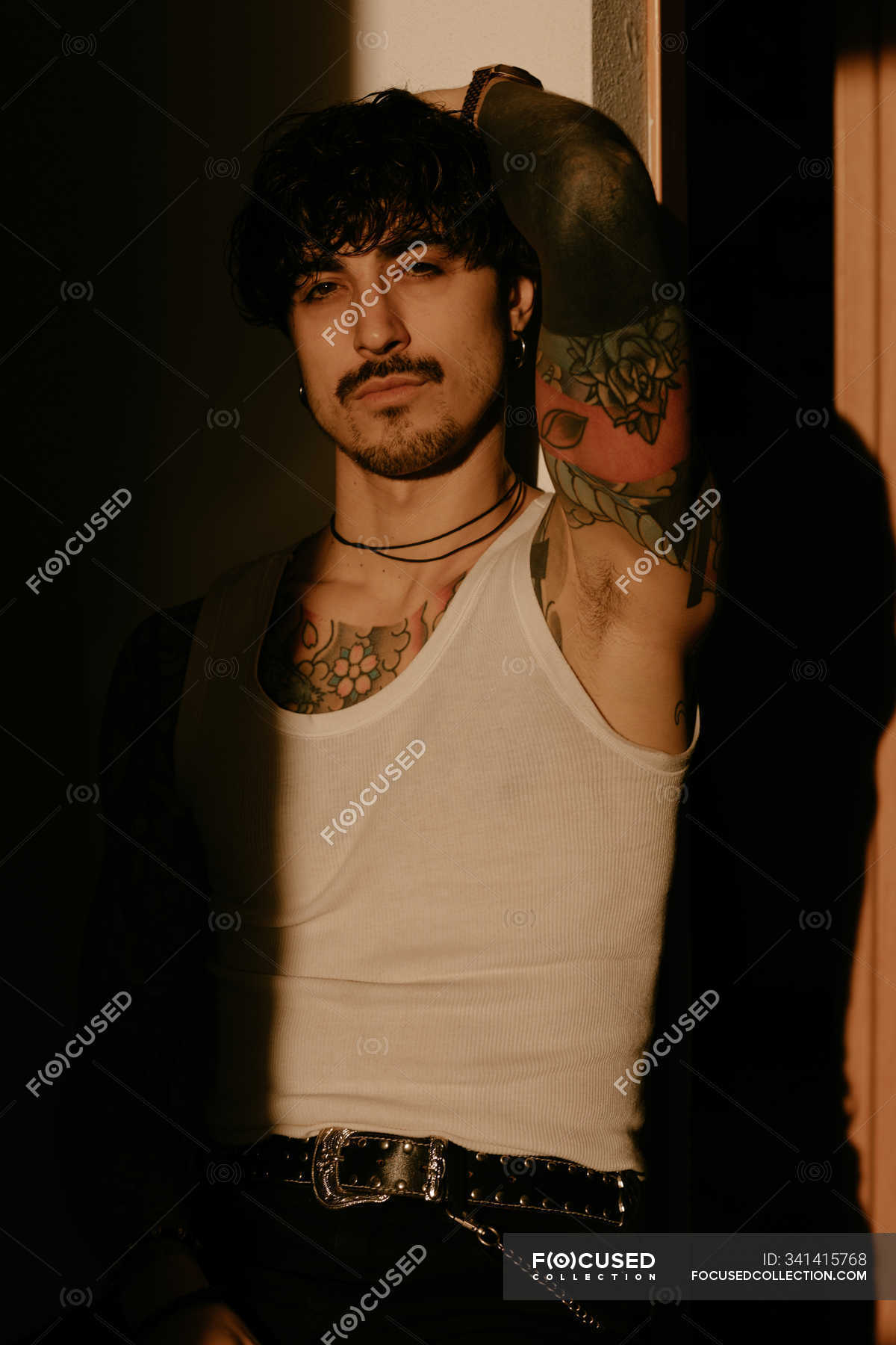 Young stylish hipster man with tattoos in white tank top leaning against  wall, looking in camera — fashionable, indoors - Stock Photo | #341415768