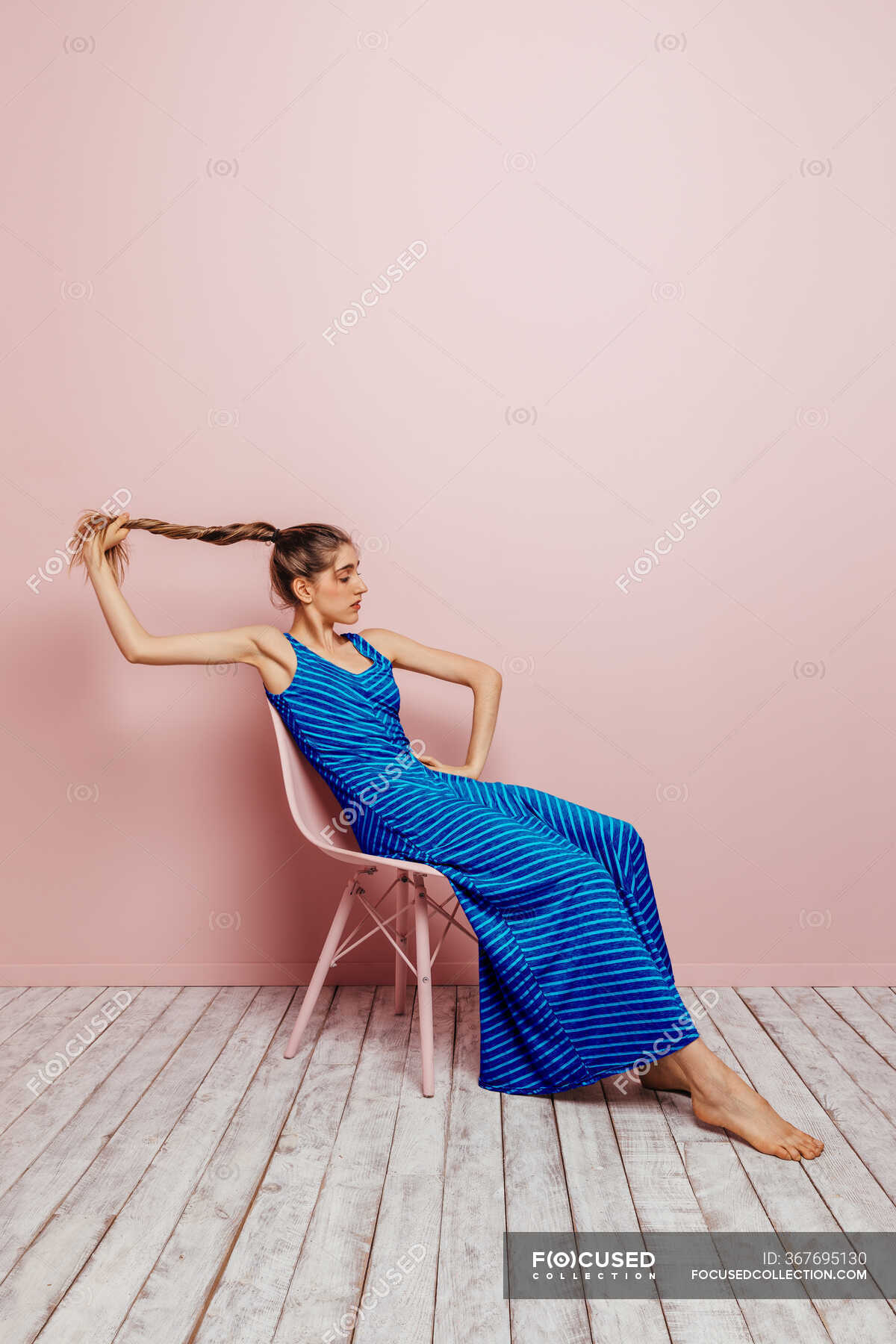 Side View Of Young Woman In Blue Dress Barefoot Looking At Camera Holding Ponytail While Sitting In Chair And Looking Away On Minimalist Pink Background Beautiful Female Stock Photo 367695130