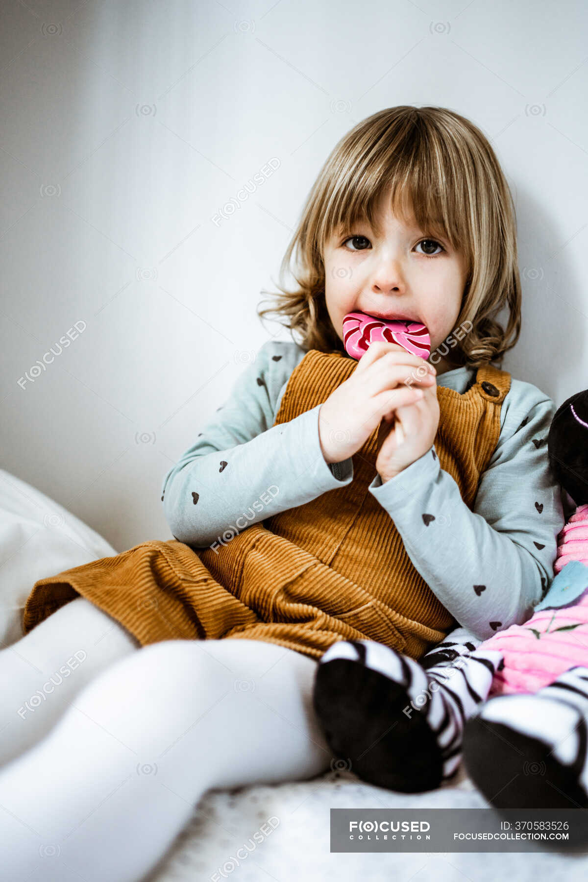 Cute little girl in casual clothes looking at camera and sucking lollipop  while sitting on comfortable bed near toy at home — joy, vertical - Stock  Photo | #370583526