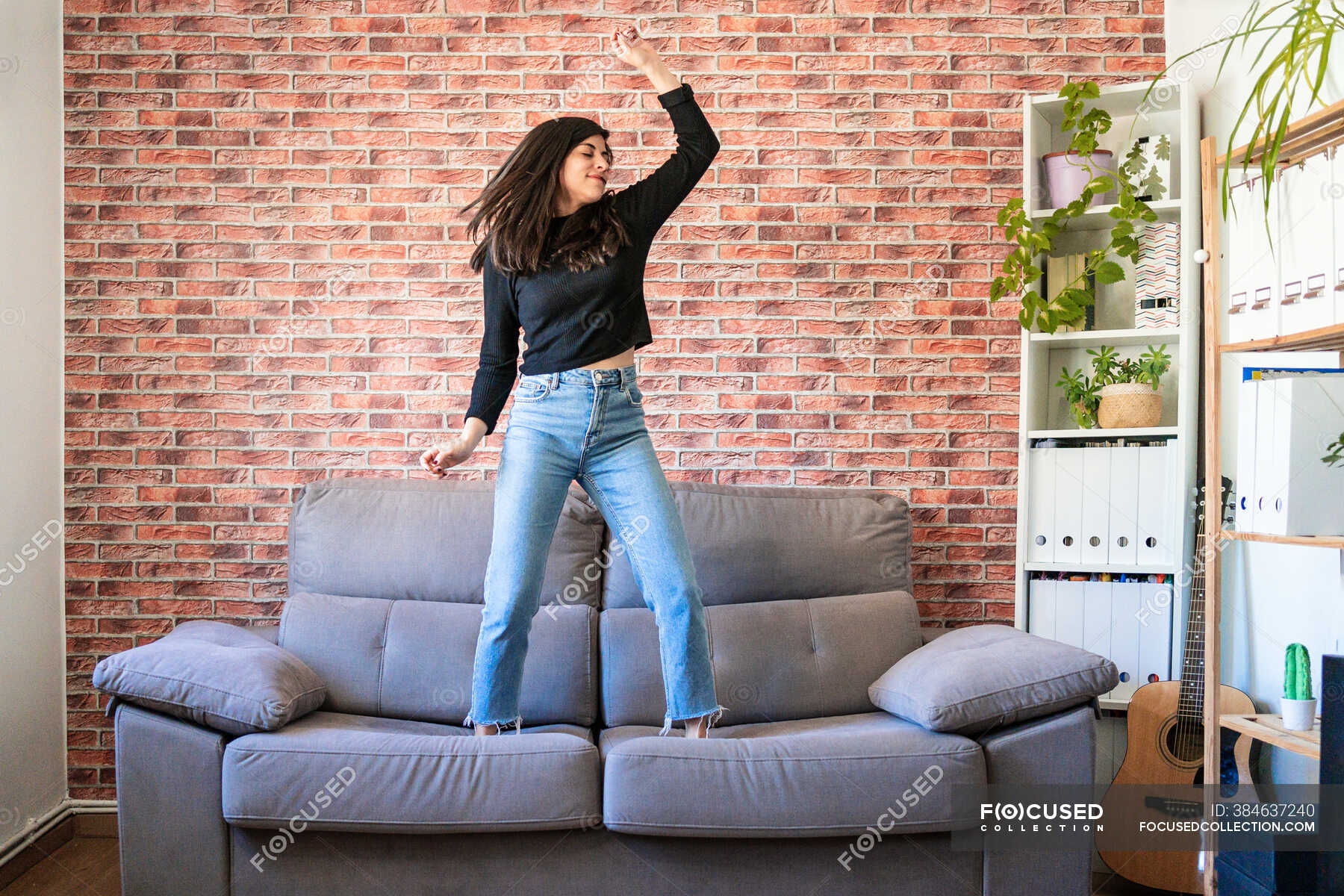 Young woman dancing on a couch in her house. Behind it is a brick wall