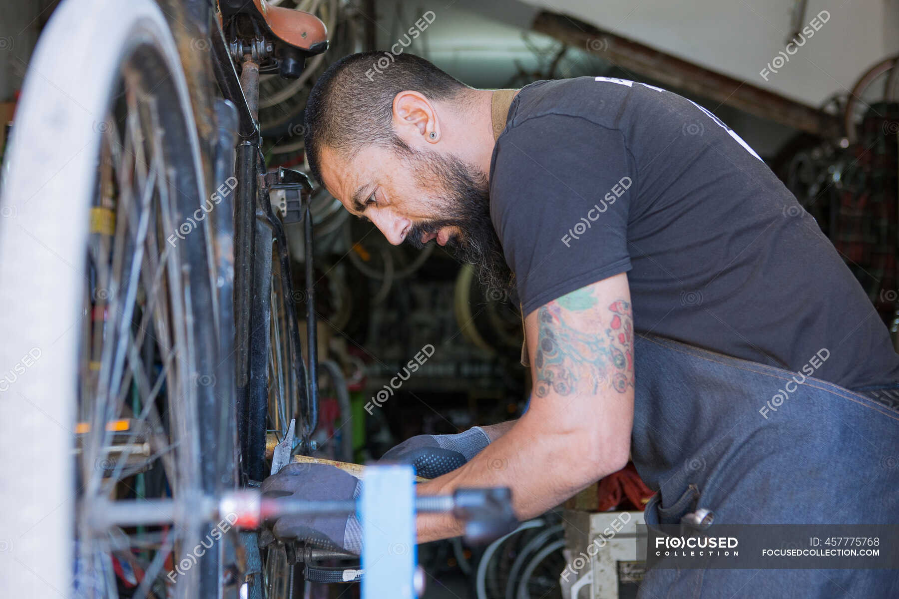 Concentrated male mechanic with beard and tattoos in gloves repairing bicycle in modern workshop — wheels, bikes - Stock Photo