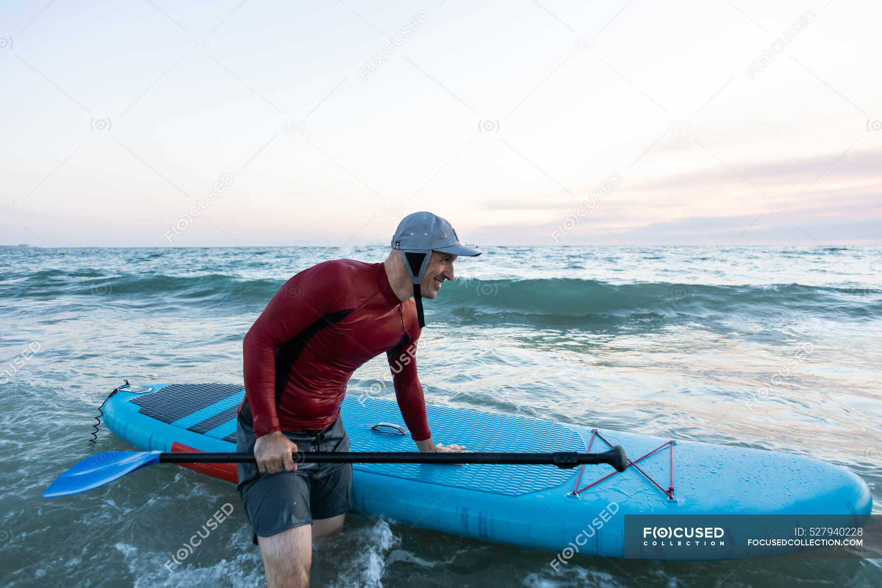 Side view of male surfer in wetsuit and hat carrying paddle board and entering water to surf on