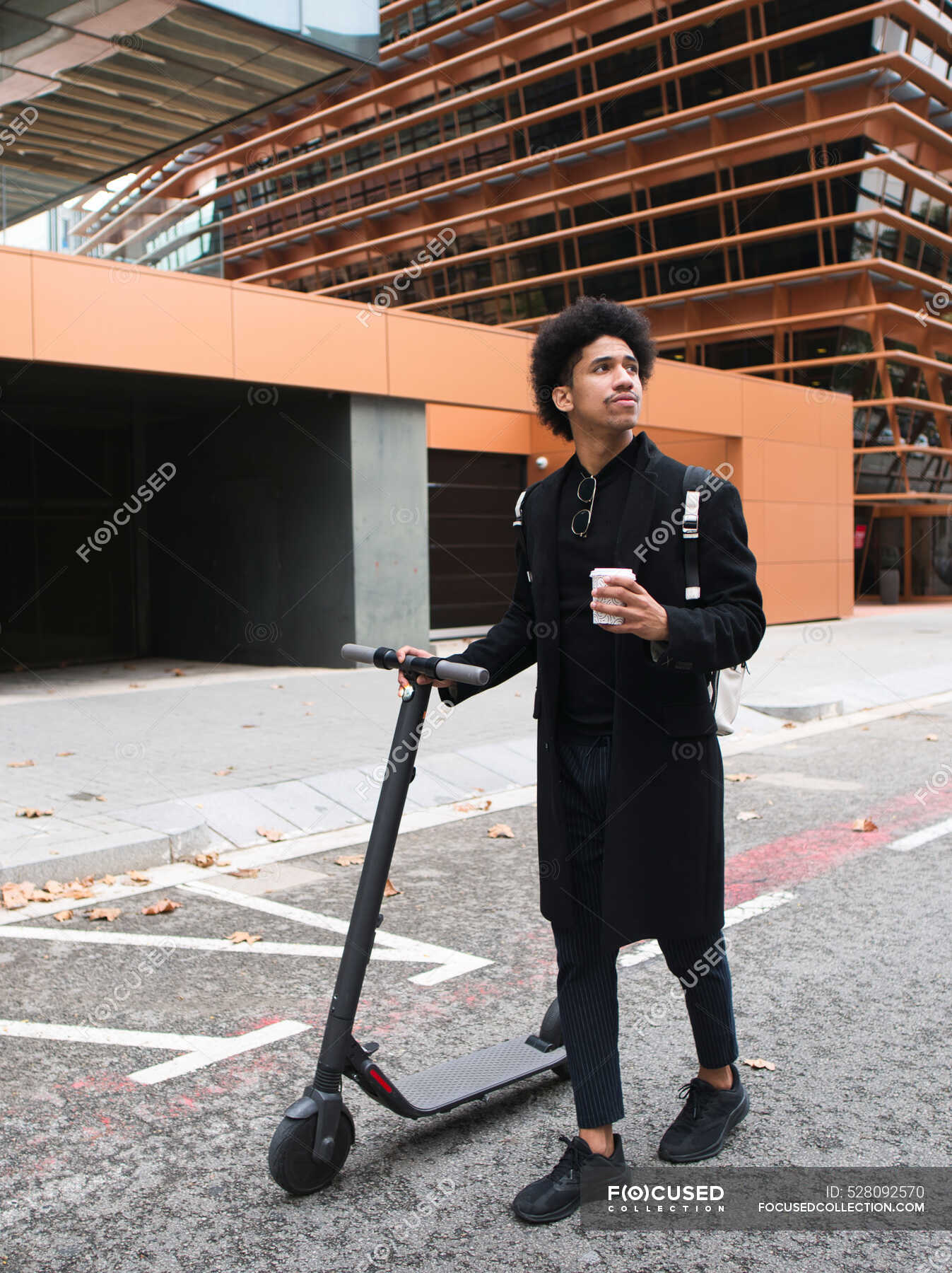 Young stylish African American male with curly dark hair in elegant coat  standing on street with electric scooter and cup of takeaway coffee and  looking away — man, to go - Stock