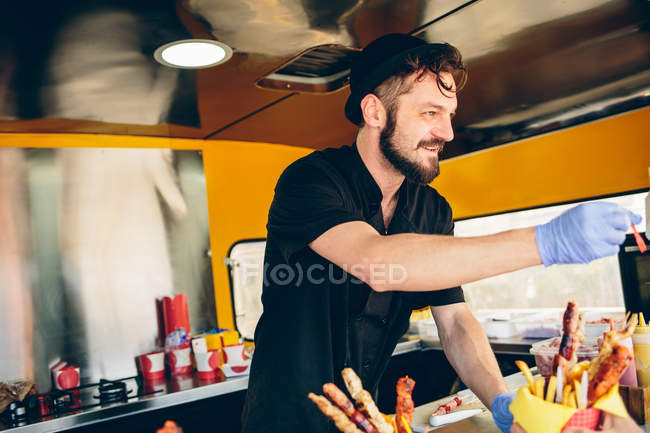 Hipster em chapéu vendendo comida em caminhão de alimentos — Fotografia de Stock