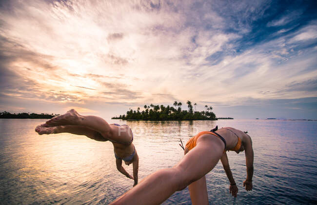 Des femmes anonymes en maillots de bain sautant dans l'eau sur fond d'île tropicale et de ciel. — Photo de stock