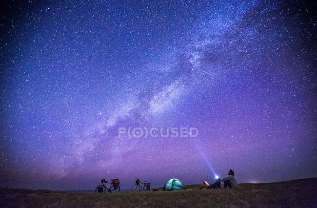 Turistas anónimos explorando numerosas estrellas en el cielo por la noche mientras están sentados en el suelo cerca de la tienda de campaña. - foto de stock