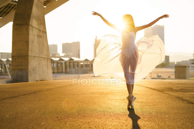 Young ballet dancer performing ballet with flying skirt and arms up against sunrise. — Stock Photo