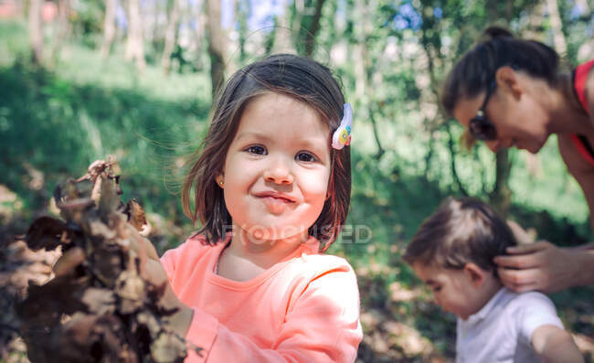 Портрет красивої маленької дівчинки, яка посміхається на камеру проти матері з сином у парку. — стокове фото