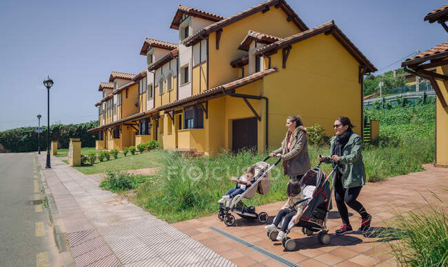 Two adult mothers pushing strollers with children in the sunny street — Stock Photo
