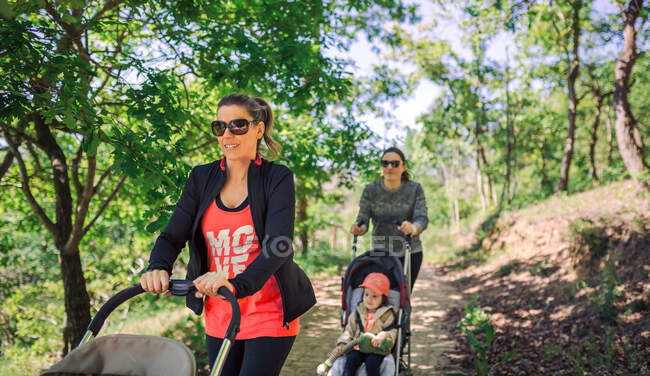 Mujeres sonrientes con gafas de sol caminando con sus bebés en cochecitos - foto de stock