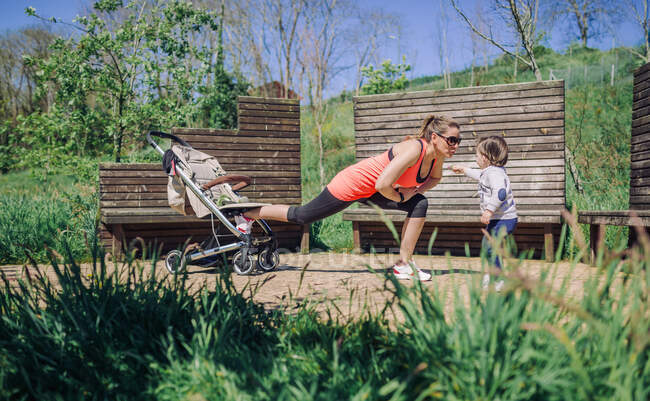 Adult mother exercising in park using baby carriage with kid beside — Stock Photo