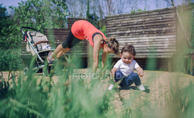 Side view of strong sporty mother doing abs workout using kid's pram while her child playing in sunlight.Grass on foreground. — Stock Photo