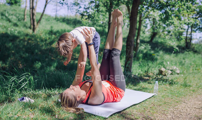 Vista della donna sportiva che esegue allenamento con il suo bambino sulle braccia sollevate con le gambe in alto nel parco — Foto stock