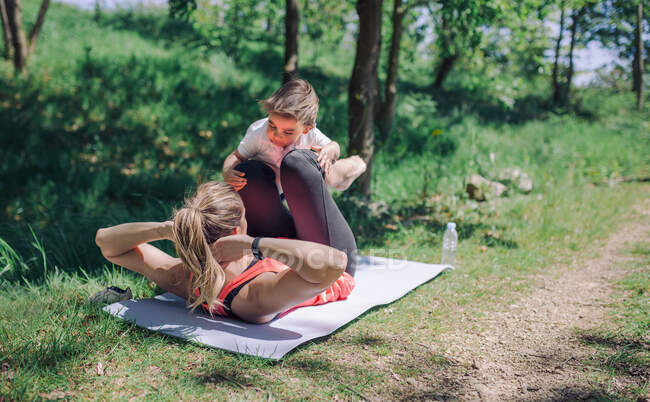Back view of strong and healthy mother doing workout in park while lying on mat with kid — Stock Photo