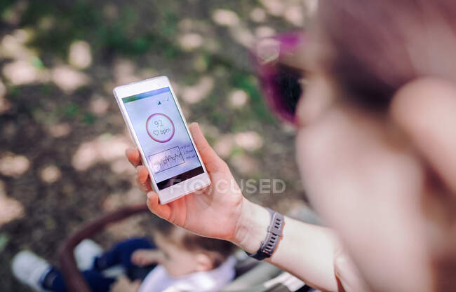 Above view of unrecognizable woman with pram in park looking at mobile screen with pulse rate.Bokeh — Stock Photo
