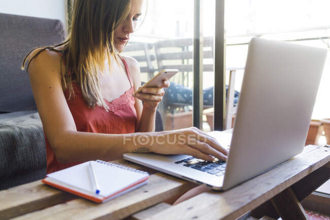 Freelancer femenina en ropa de casa trabajando en el ordenador portátil y el uso de teléfono móvil en la sala de estar - foto de stock