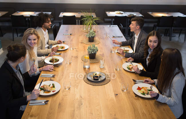 Group of young coworkers having dinner together sitting at big table in spacious light canteen. — Stock Photo