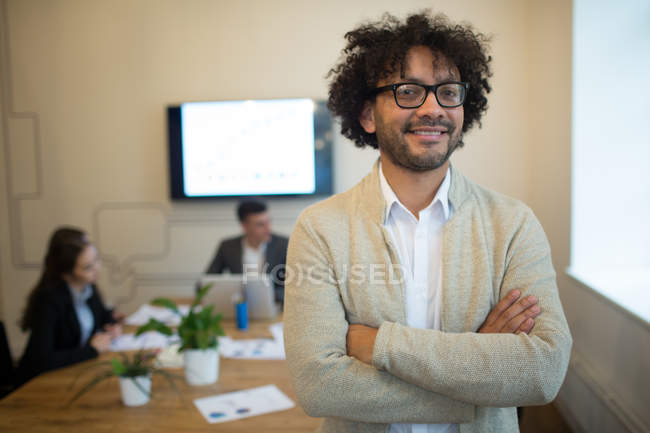 Lächelnder Mann mit Brille, die Hände über dem Kopf verschränkt und vor dem Hintergrund des Büros in die Kamera blickend — Stockfoto