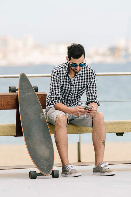 Молодой человек сидит на скамейке со смартфоном — стоковое фото