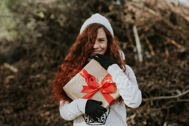 Porträt eines lächelnden Ingwermädchens mit Strickmütze, das ein schön verpacktes Geschenk in der Hand hält — Stockfoto