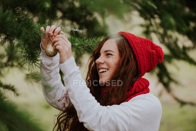 Portrait of laughing ginger girl in knitted red hat decorating fir tree outside — Stock Photo