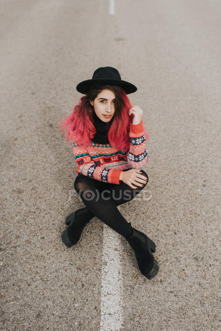 Девушка с розовыми волосами в шляпе сидит на дороге — стоковое фото