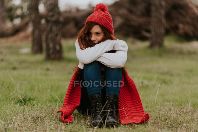 Portrait of young pretty girl in red knitted heading and scarf sitting on ground against blurred forest. — Stock Photo