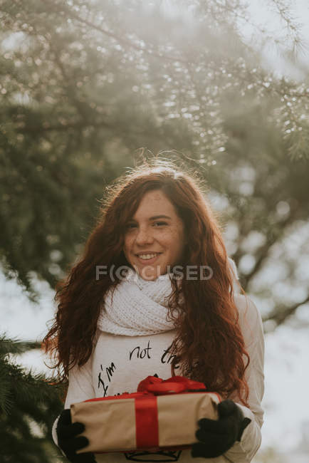 Portrait of a smiling ginger girl holding present looking at camera in winter forest — Stock Photo