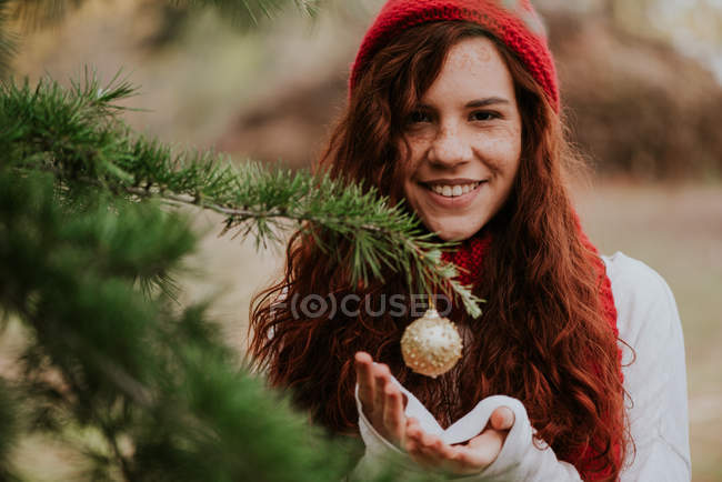 Portrait of smiling redhead girl holding hands under bauble hanging on pine tree. — Stock Photo