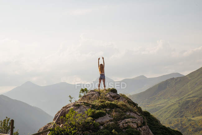 Rear view of blond girl posing with raised hands on mountain peak at highlands — Stock Photo