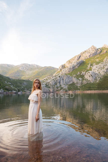 Portrait of blonde girl wearing white dress standing in mountain lake and looking over shoulder away — Stock Photo