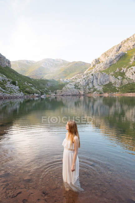Blonde girl in white dress posing in mountain lake. — Stock Photo