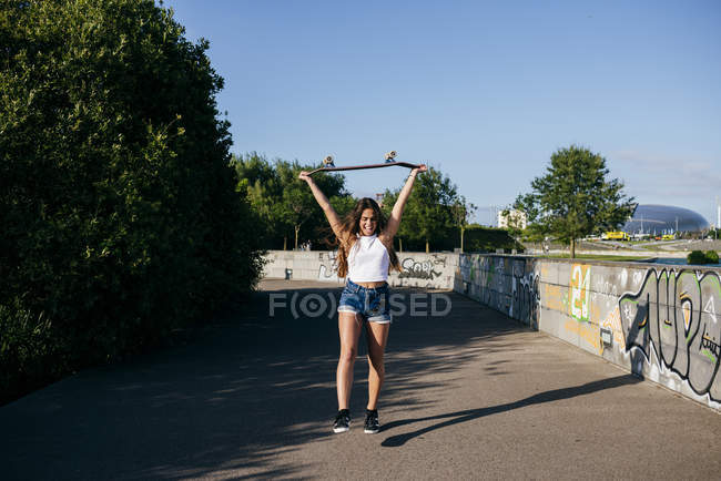 Menina feliz segurando skate acima da cabeça — Fotografia de Stock