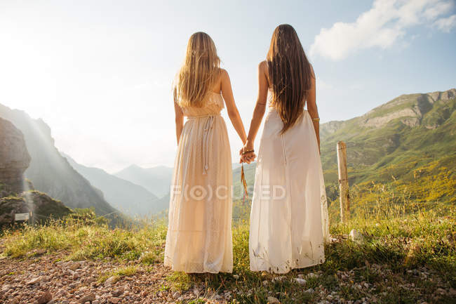 Вид сзади на двух девушек в белом платье, держащихся за руки и позирующих в горной местности — стоковое фото